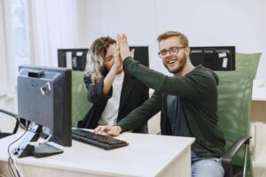 Two coworkers in modern office giving a high five in front of computer screens, symbolizing teamwork.